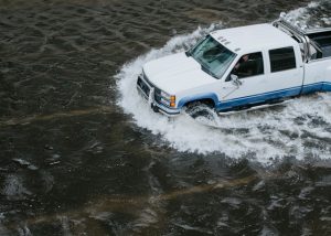 White pickup truck driving through high floodwater, demonstrating the risks of vehicles in floods.