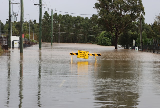 Street covered in floodwater with a caution sign partly submerged, highlighting dangerous road conditions.