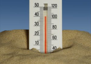 Thermometer placed in hot sand under a clear blue sky, symbolizing extreme heat and rising temperatures.